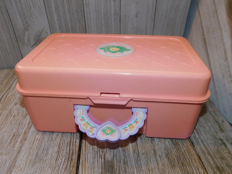 Puede incluir: Una caja de almacenamiento de pl&aacute;stico rosa con un asa morada y un dise&ntilde;o de flor en la tapa. La caja tiene un cierre en la parte delantera y una peque&ntilde;a asa en la parte superior.