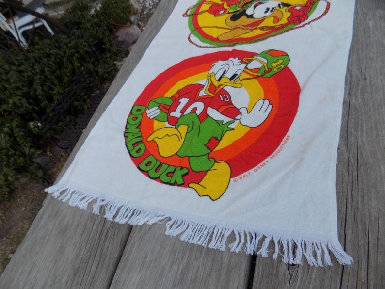 Peut inclure: Serviette en coton blanc avec un dessin color&eacute; de Donald Duck en uniforme de football. Le motif comprend un fond circulaire rouge, orange et jaune avec les mots "DONALD DUCK". La serviette a une bordure &agrave; franges.