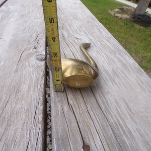 Pu&ograve; includere: Un oggetto decorativo a forma di cigno in ottone, che misura circa 13 cm di lunghezza, &egrave; mostrato su una superficie di legno.