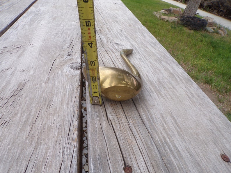 Pu&ograve; includere: Un oggetto decorativo a forma di cigno in ottone, che misura circa 13 cm di lunghezza, &egrave; mostrato su una superficie di legno.