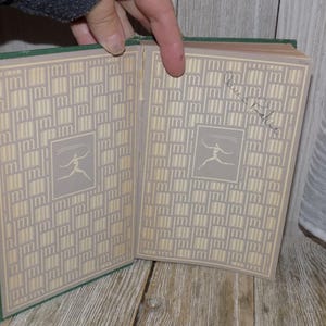 May include: A green hardcover book with a cream-colored patterned cover. The book is open to reveal a cream-colored page with a stylized illustration of a person.