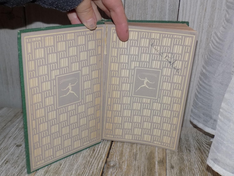 May include: A green hardcover book with a cream-colored patterned cover. The book is open to reveal a cream-colored page with a stylized illustration of a person.