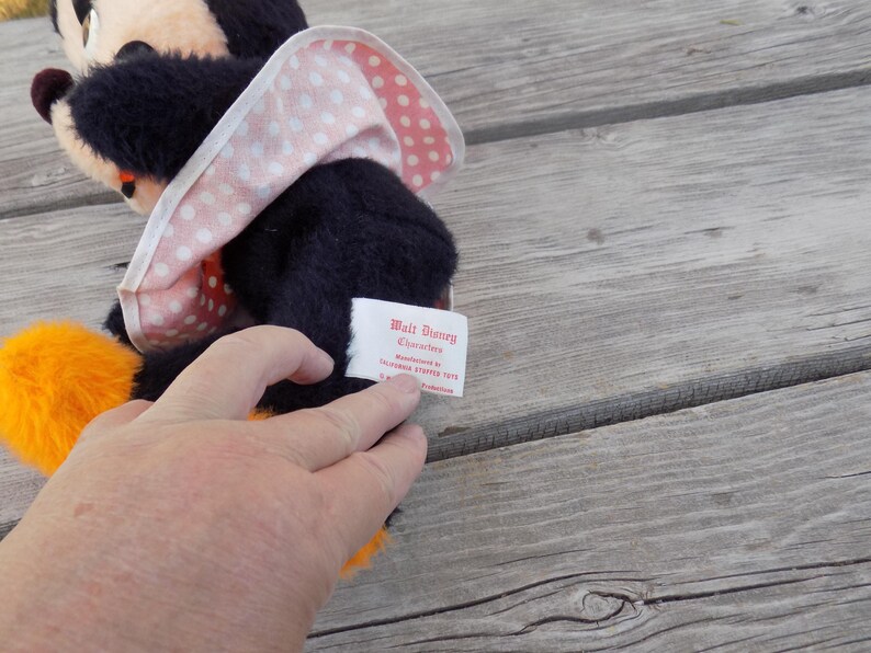May include: A vintage Minnie Mouse plush toy with a black body, orange feet, and a pink and white polka dot bow. The tag reads "Walt Disney Characters". The plush is on a weathered wooden surface.