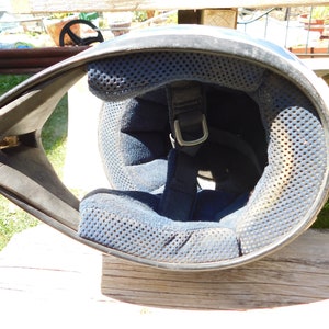 May include: A white motorcycle helmet with a black chin guard and a blue padded interior. The helmet has a strap with a metal buckle.