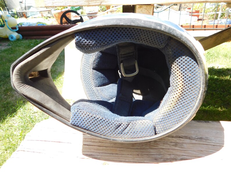 May include: A white motorcycle helmet with a black chin guard and a blue padded interior. The helmet has a strap with a metal buckle.