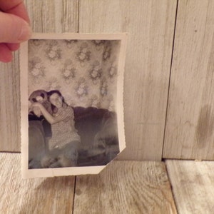 May include: A monochrome photograph depicts a person hugging a teddy bear on a sofa. The image, with a torn edge, is held against a wooden backdrop. The person is wearing a checked shirt.