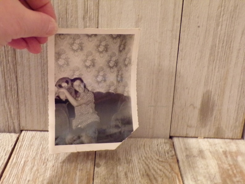 May include: A monochrome photograph depicts a person hugging a teddy bear on a sofa. The image, with a torn edge, is held against a wooden backdrop. The person is wearing a checked shirt.