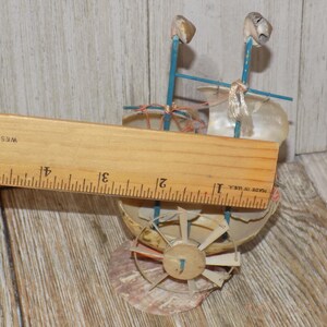 May include: A miniature seashell boat with blue supports and a seashell sail. The boat has a wooden wheel and is approximately 4 inches long, as measured by a wooden ruler. The boat is made of seashells and other natural materials.