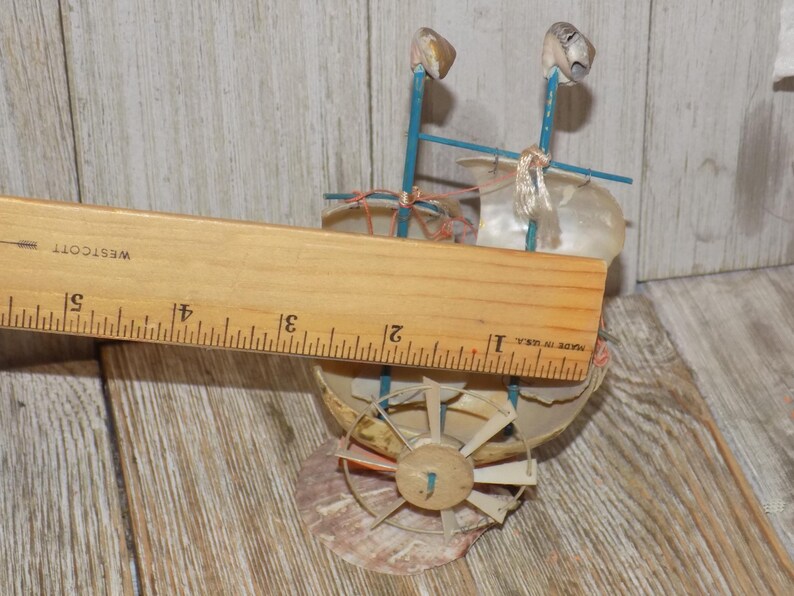 May include: A miniature seashell boat with blue supports and a seashell sail. The boat has a wooden wheel and is approximately 4 inches long, as measured by a wooden ruler. The boat is made of seashells and other natural materials.