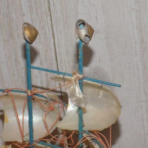 May include: A miniature sailboat model with a blue frame, adorned with shell sails and gold-tipped masts. The sails are made of translucent material, and the rigging is a reddish-orange color. A decorative nautical item.