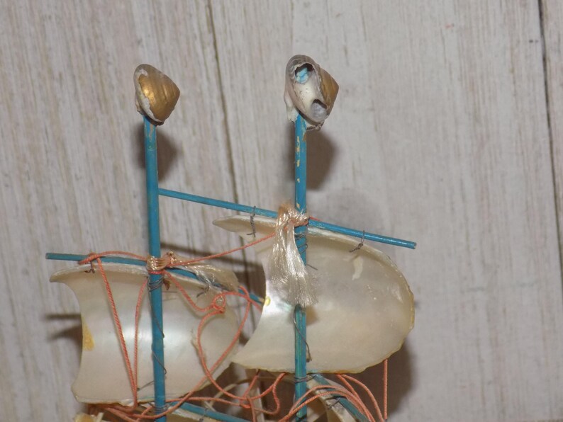 May include: A miniature sailboat model with a blue frame, adorned with shell sails and gold-tipped masts. The sails are made of translucent material, and the rigging is a reddish-orange color. A decorative nautical item.