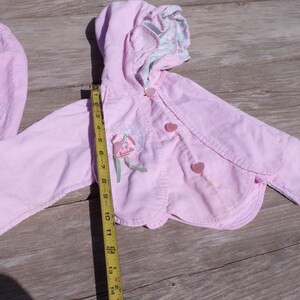Puede incluir: Una chaqueta de pana rosa con capucha y bordado floral. La chaqueta tiene dos botones en forma de coraz&oacute;n y un dobladillo festoneado. La chaqueta se mide con una cinta m&eacute;trica.