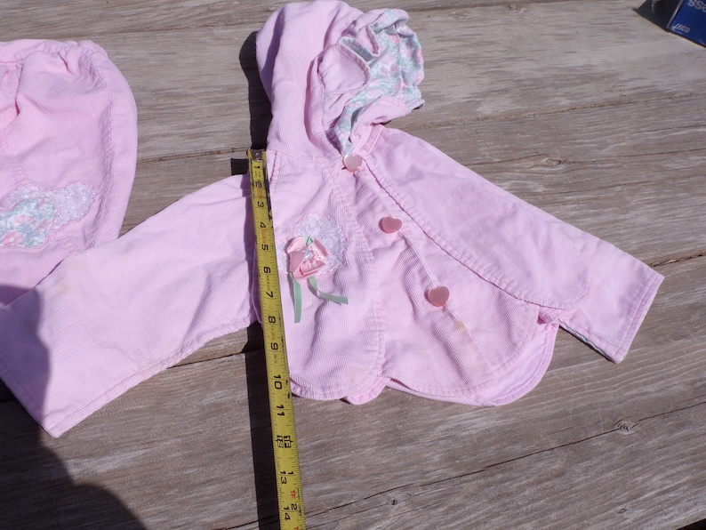 Puede incluir: Una chaqueta de pana rosa con capucha y bordado floral. La chaqueta tiene dos botones en forma de coraz&oacute;n y un dobladillo festoneado. La chaqueta se mide con una cinta m&eacute;trica.