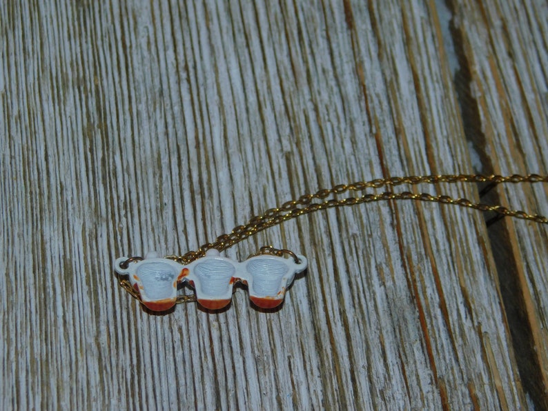 May include: A gold chain necklace with a pendant featuring three miniature teacups with white interiors and red and orange accents.
