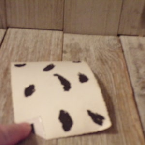 May include: A piece of off-white paper with irregular black spots, resembling a Dalmatian pattern. The paper is slightly curved and appears to be folded at one corner. The background is a light-coloured wooden surface.