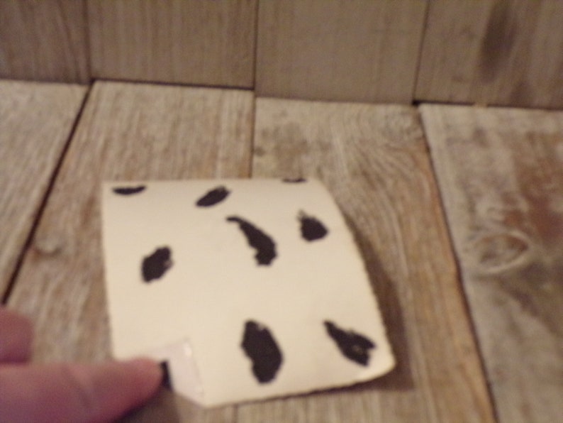 May include: A piece of off-white paper with irregular black spots, resembling a Dalmatian pattern. The paper is slightly curved and appears to be folded at one corner. The background is a light-coloured wooden surface.
