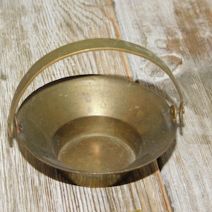 May include: A small, brass bowl with a handle. The bowl is round and has a smooth, shiny surface. It is sitting on a wooden surface.