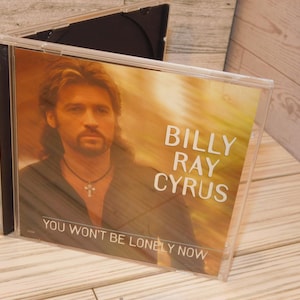 May include: A CD case featuring the album "You Won't Be Lonely Now" by Billy Ray Cyrus. The cover art shows a portrait of Cyrus with long hair and a necklace. The background has a warm, golden hue.