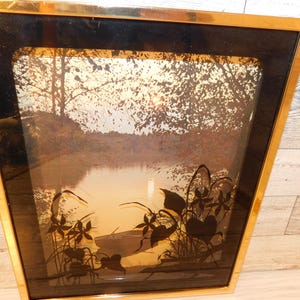 May include: Framed landscape art featuring a serene lake scene with trees and a sunset. The artwork is encased in a gold-toned frame with a black inner border. Silhouetted plants are in the foreground.