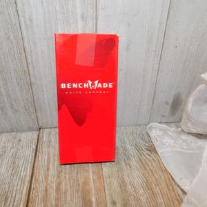 May include: A bright red box with the "Benchmade Knife Company" logo in white. The box is rectangular and stands upright on a wooden surface. The background is a light wood grain.