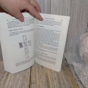 May include: An open book with a white background. The book is open to a page with the title "Card Cross Stunt" and a diagram of nine playing cards arranged in a cross shape. The next page has the title "24 Stunts with Cards" and text describing a card trick.