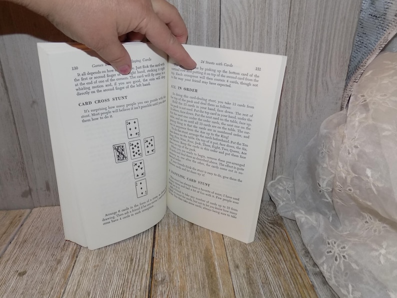 May include: An open book with a white background. The book is open to a page with the title "Card Cross Stunt" and a diagram of nine playing cards arranged in a cross shape. The next page has the title "24 Stunts with Cards" and text describing a card trick.