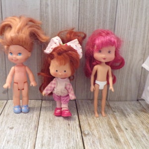 Pode incluir: Tr&ecirc;s bonecas Strawberry Shortcake com cabelo ruivo. As bonecas est&atilde;o vestindo roupas diferentes. A boneca &agrave; esquerda est&aacute; usando sapatos azuis. A boneca do meio est&aacute; usando uma camisa xadrez rosa e branca e cal&ccedil;as rosa. A boneca &agrave; direita est&aacute; usando roupa interior branca.