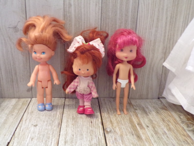 Pode incluir: Tr&ecirc;s bonecas Strawberry Shortcake com cabelo ruivo. As bonecas est&atilde;o vestindo roupas diferentes. A boneca &agrave; esquerda est&aacute; usando sapatos azuis. A boneca do meio est&aacute; usando uma camisa xadrez rosa e branca e cal&ccedil;as rosa. A boneca &agrave; direita est&aacute; usando roupa interior branca.