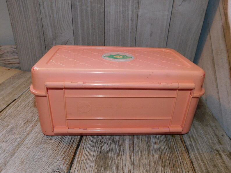 Puede incluir: Una caja de almacenamiento de pl&aacute;stico rosa con una tapa con cierre. La tapa tiene una pegatina verde y blanca con un personaje de dibujos animados.