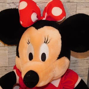 Op de afbeelding: Een pluche Minnie Mouse pop met grote zwarte oren en een rode strik met witte stippen. De pop heeft een rode jurk en een zwarte neus. Het gezicht is licht perzik met geborduurde ogen en wimpers.