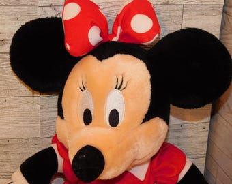 May include: A plush Minnie Mouse doll with large black ears and a red bow with white polka dots. The doll has a red dress and a black nose. The face is light peach with embroidered eyes and eyelashes.