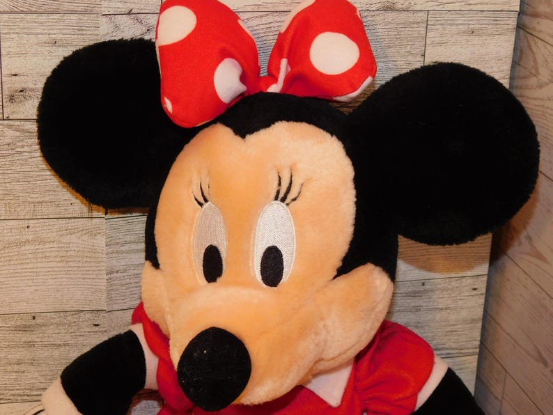 Op de afbeelding: Een pluche Minnie Mouse pop met grote zwarte oren en een rode strik met witte stippen. De pop heeft een rode jurk en een zwarte neus. Het gezicht is licht perzik met geborduurde ogen en wimpers.