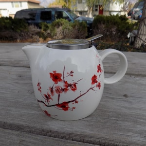 May include: White ceramic teapot with a stainless steel lid and a floral design of red cherry blossoms on a branch. The teapot has a handle and a spout.