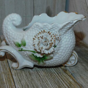 May include: White porcelain planter shaped like a carriage with a white flower and green leaves. The planter has a textured surface and gold accents.