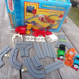 May include: A Thomas & Friends Big Loader toy train set with a green and orange train engine, a blue train engine, and a gray track. The set includes a red and white loading station, a yellow sign, and a white fence.