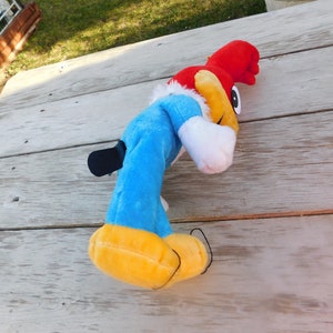 May include: A blue and red plush toy of Woody Woodpecker, a cartoon character. The toy is lying on a wooden surface.