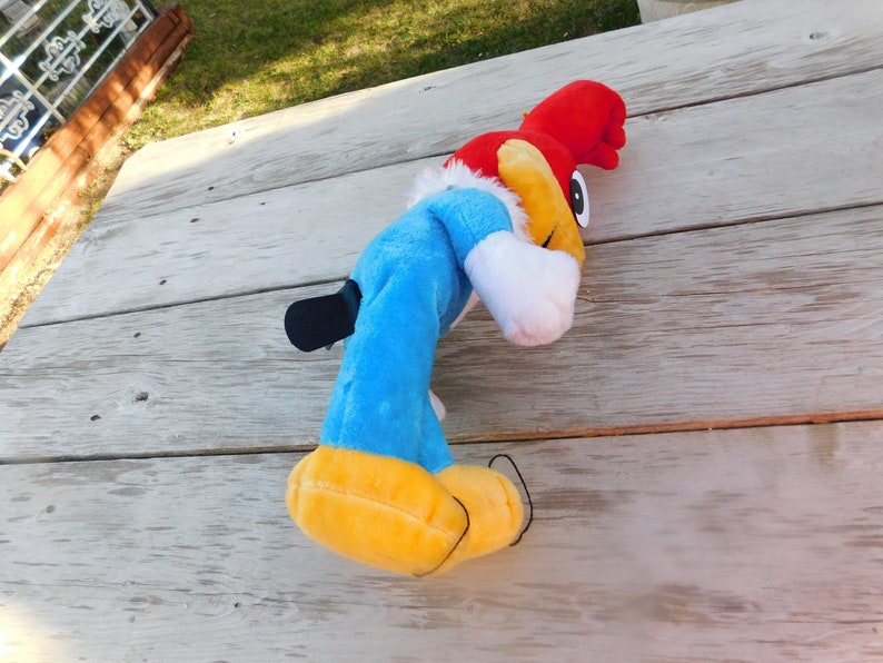 May include: A blue and red plush toy of Woody Woodpecker, a cartoon character. The toy is lying on a wooden surface.