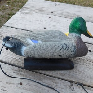 May include: A green-headed, gray and brown duck decoy with a black base and a black cord attached. The decoy is sitting on a wooden surface.