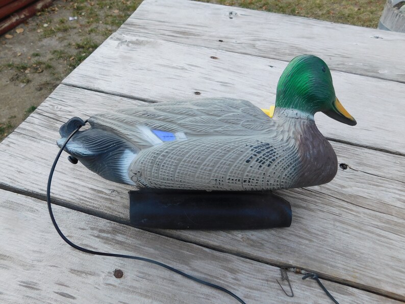 May include: A green-headed, gray and brown duck decoy with a black base and a black cord attached. The decoy is sitting on a wooden surface.