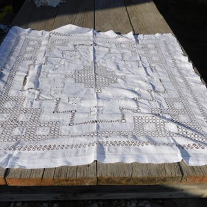 May include: A white, rectangular tablecloth with intricate cutwork and a scalloped edge. The detailed openwork design features geometric patterns and floral motifs. The tablecloth is displayed on a weathered wooden surface, showcasing its delicate craftsmanship.