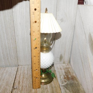 May include: A white milk glass oil lamp with a gold base and a white fabric lampshade. The lamp is about 7 inches tall and has a green leaf design on the base.