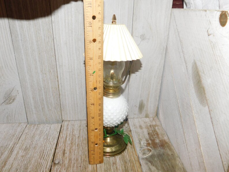 May include: A white milk glass oil lamp with a gold base and a white fabric lampshade. The lamp is about 7 inches tall and has a green leaf design on the base.