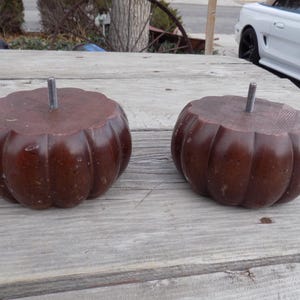 May include: Two dark brown, pumpkin-shaped wooden furniture legs with a threaded metal rod protruding from the top. The legs are sitting on a weathered wooden surface.