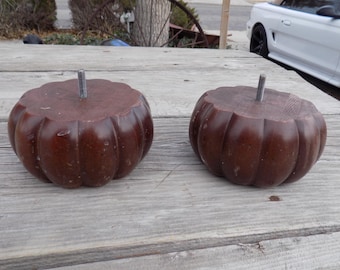 May include: Two dark brown, pumpkin-shaped wooden furniture legs with a threaded metal rod protruding from the top. The legs are sitting on a weathered wooden surface.