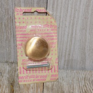 May include: A new, round, gold-colored cabinet knob with a screw, packaged on a pink and yellow card. The card has the text "brand merit" and "MADE IN U.S.A. BY AMEROCK".