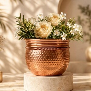 Puede incluir: Una maceta redonda de cobre martillado con un detalle decorativo de borde estilo cuerda. La maceta está llena de un ramo de rosas color crema y pequeñas flores blancas, creando un arreglo floral brillante en un recipiente metálico.