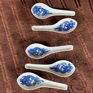 May include: Five white ceramic soup spoons arranged in a vertical column. Each spoon features a blue underglaze design of a dragon with swirling patterns. The spoons have a wide, rounded bowl and a short, straight handle, all made of porcelain.