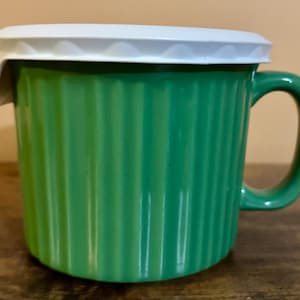 May include: A green mug with a vertical ribbed texture and a matching green handle. The mug is topped with a white plastic lid that features a scalloped edge design. The vessel has a cylindrical shape and a smooth, glossy finish on the exterior surface.
