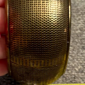 May include: A wide, cylindrical brass bangle bracelet with a fine, textured mesh pattern across its surface. A yellow measuring tape is positioned at the base, indicating the bracelet has a width of approximately 1.5 inches.