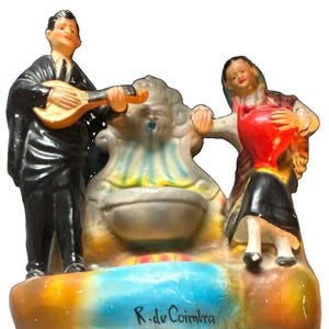 May include: A ceramic figurine featuring a person in a black suit playing a lute and another person holding a red pitcher, both standing beside a central fountain. The base is painted in blue and orange with the text R. de Coimbra written in black.